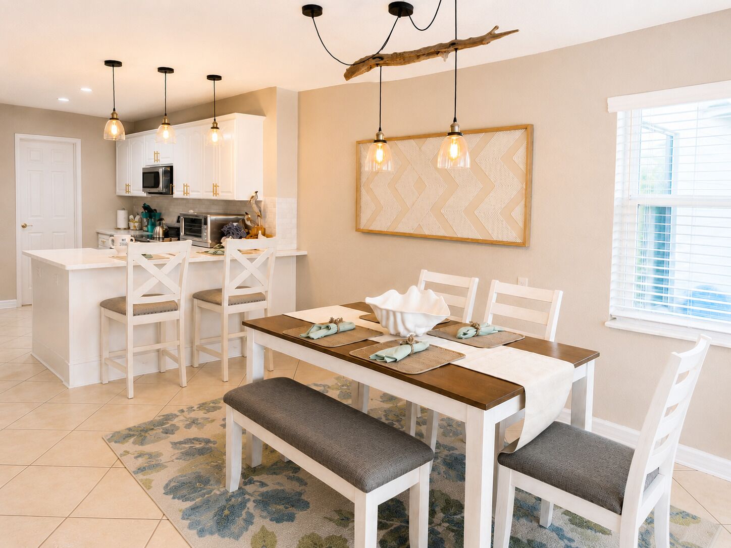 Un coin repas avec une table blanche, des chaises et un banc. Des lampes suspendues sont placées au-dessus de la table.