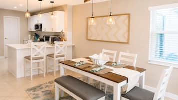 Un coin repas avec une table blanche, des chaises et un banc. Des lampes suspendues sont placées au-dessus de la table.