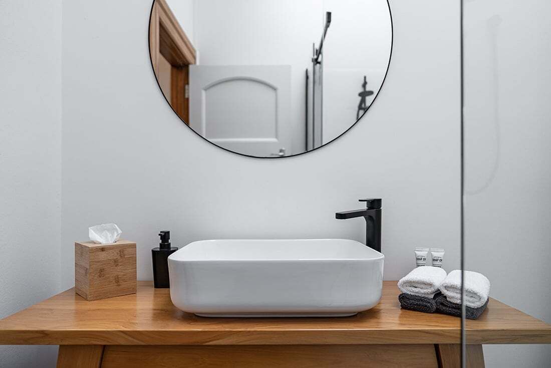 Une salle de bain avec un lavabo blanc, un robinet noir et un miroir rond. On peut voir une boîte en bois et une bouteille sur le comptoir.