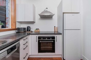 A compact kitchen with white cabinetry, a built-in oven, a stainless steel range hood, and a white refrigerator. - Esplanade Apartments Seashells (Dunedin)
