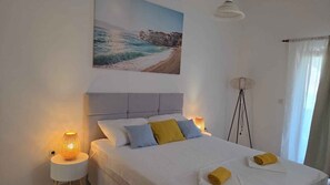 Un dormitorio con cama, dos lámparas de noche, un cuadro de una escena costera, una lámpara de pie y una ventana con cortinas.