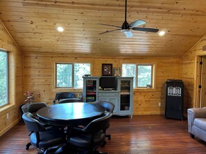 A wooden cabin interior with a dining table, chairs, a ceiling fan, a television, and a cabinet with books. - Terrific 3-bedroom cabin with AC in cozy Fish Camp (Fish Camp)