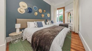 A bedroom with a bed, bedside tables, a wall-mounted hat display, and a window with curtains. - Esplanade Apartments Wavecaps (Dunedin)