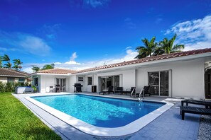 Ein Pool mit Fliesen umgeben, Sitzgelegenheiten im Freien und ein Gebäude mit blauem Flachdach.