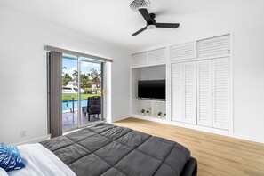 Une chambre avec un lit, un ventilateur de plafond, une télévision et une porte-fenêtre coulissante donnant sur un espace extérieur avec une piscine.