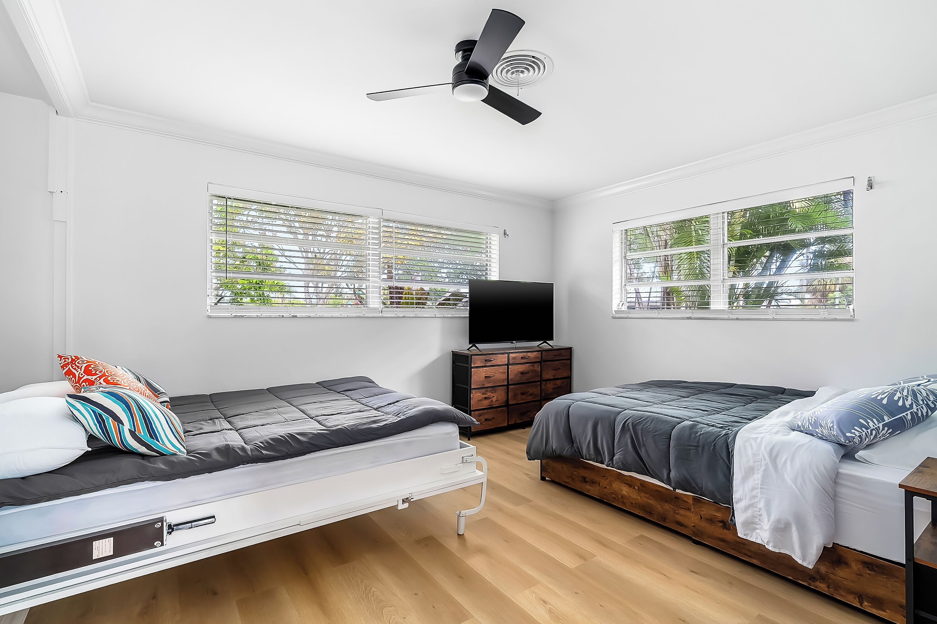 Un dormitorio con dos camas, un ventilador de techo, un televisor y ventanas amplias.