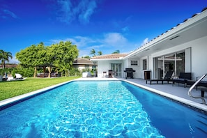 A swimming pool with a clear blue water, surrounded by a well-maintained lawn and a building with large windows and outdoor seating.