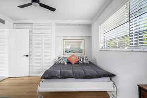 Une chambre à coucher avec un lit, un ventilateur de plafond, une fenêtre avec des stores, une porte et un tableau encadré au mur.