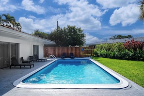 Une piscine rectangulaire avec une terrasse carrelée, entourée d’une clôture en bois, et une zone herbeuse avec une clôture en bois en arrière-plan.