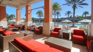 A covered outdoor seating area with wicker furniture and red cushions, overlooking a pool and palm trees. - Disney, Resort Style, 4 BDRM, Sleeps 8, 3 Bath (Kissimmee)