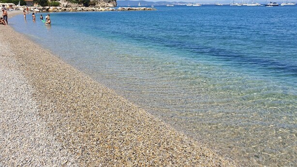 Una spiaggia di ciottoli con acqua cristallina, barche all'orizzonte e edifici su una collina.