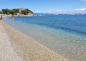 A pebble beach with clear water, boats in the distance, and buildings on a hill. - Appartamenti Il Dormentorio - Dormentorio - Bilocale PT Lato Rio (Rio Marina)