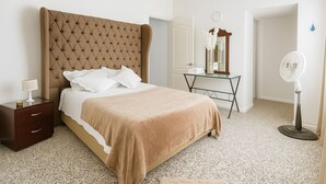 A bedroom with a tufted headboard, a bed with white and beige bedding, a nightstand with a lamp, a mirror, a desk with a fan, and a carpeted floor. - CASA LUCY ALOJAMIENTOS (Palmira)