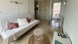 A cozy living room with a sofa, a round wicker table, and a dining area with a chair. - Studio with terrace for 2 people in Giens – Secure residence near the beaches (HYERES)
