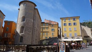 A historic tower with a clock, surrounded by colorful buildings and outdoor seating areas. - Studio with terrace for 2 people in Giens – Secure residence near the beaches (HYERES)