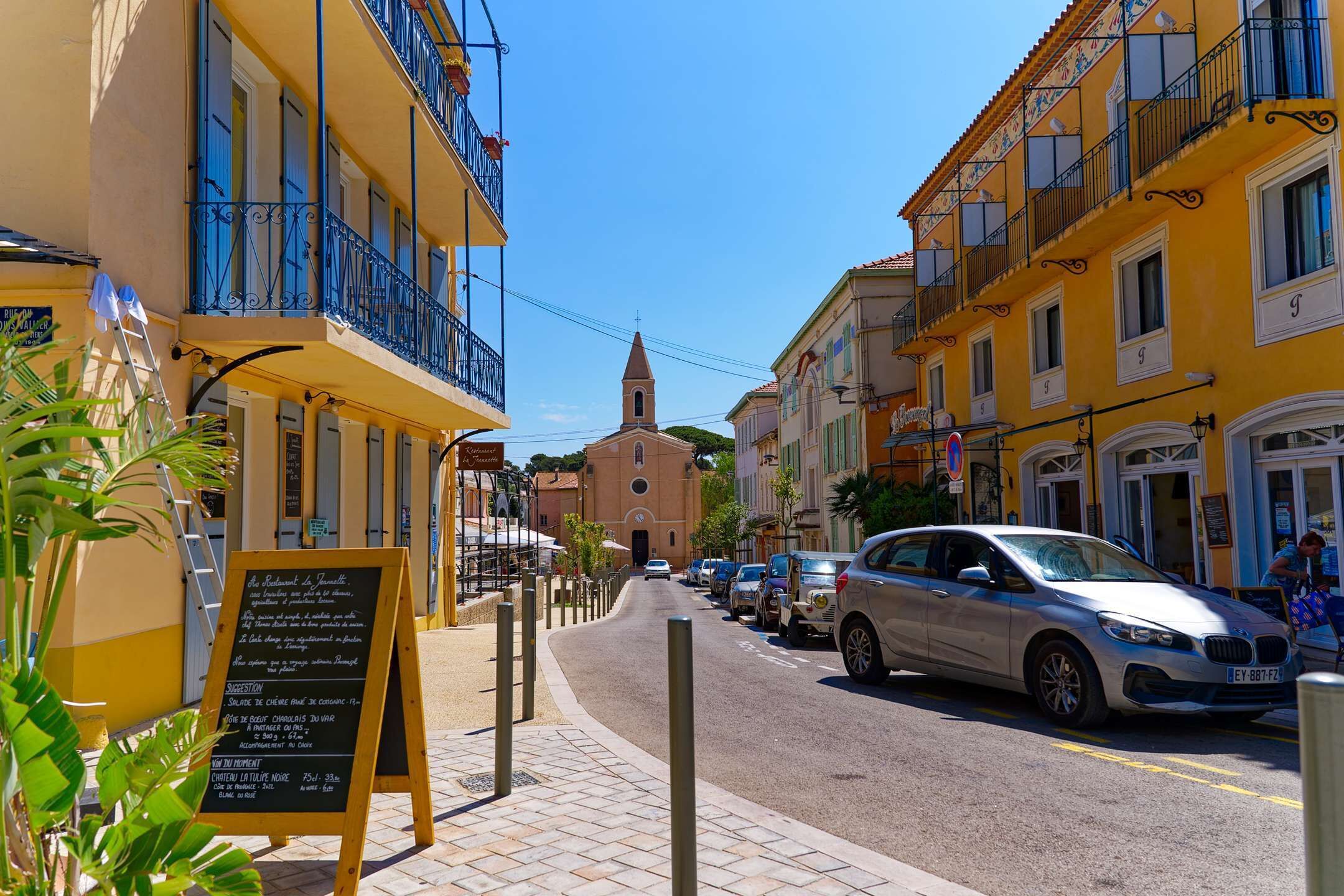 A street with yellow buildings, a church, parked cars, and a signboard.