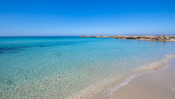 Ein sonniger Strand mit ruhigem Wasser und Felsen in der Ferne.