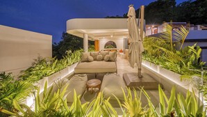 Uma área externa moderna com lounge, sofás, almofadas e uma mesa, rodeada por vegetação exuberante.