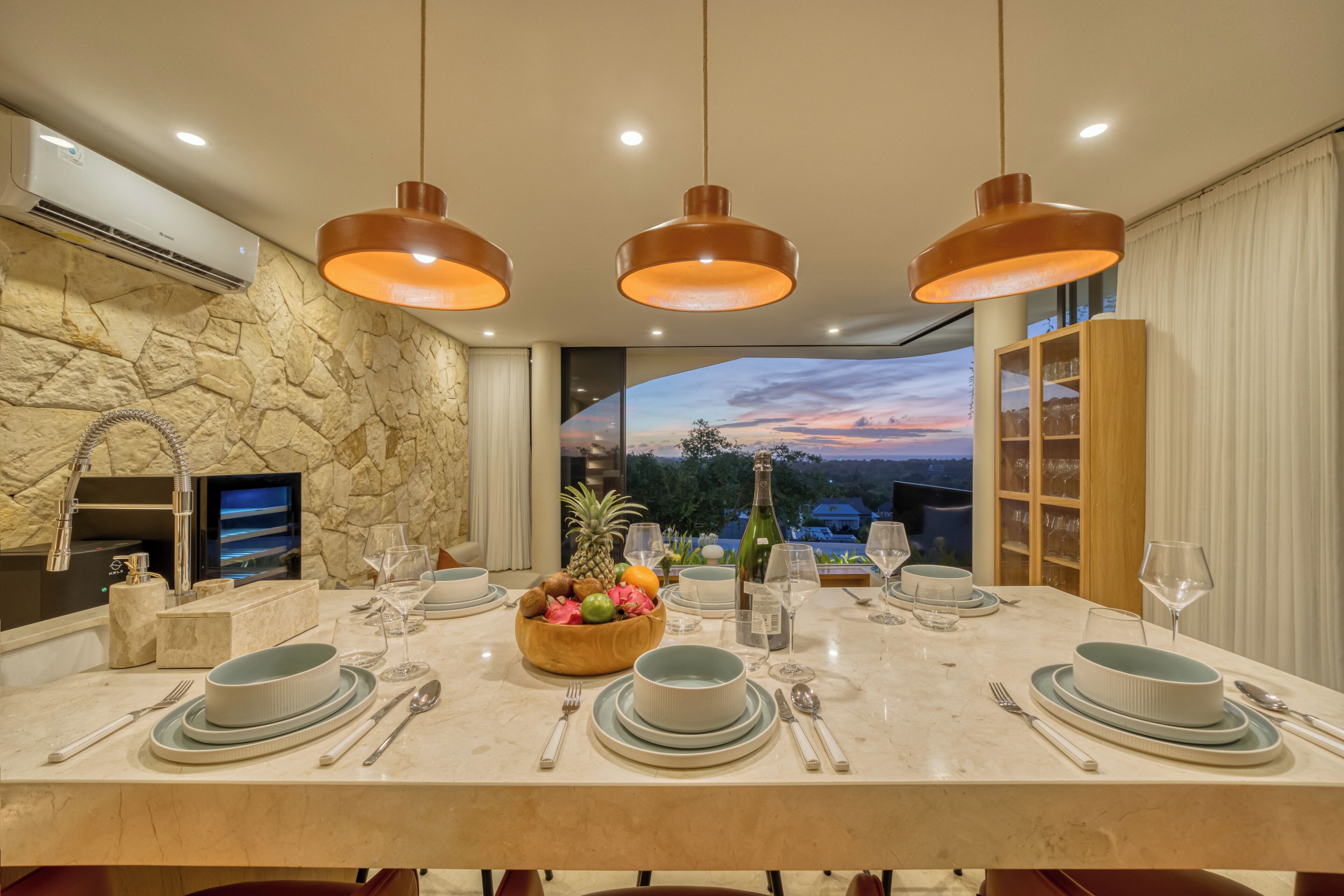 A dining area with a stone wall, a view of the sunset, and a table set with plates, glasses, and a fruit bowl.