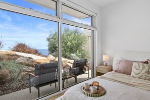 A bedroom with a large window, a bed with pillows, a bedside table with a lamp, and a tray with two mugs.