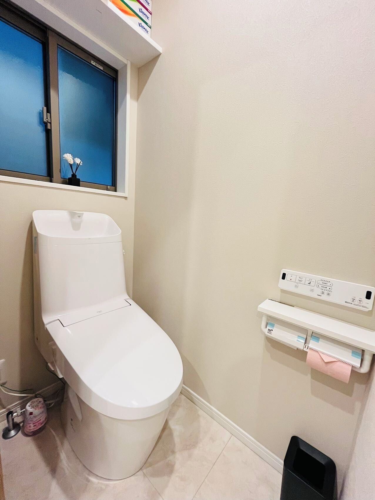 Eine weiße Toilette mit Bedienfeld, ein Fenster mit blauen Jalousien und eine kleine Ablage mit Gegenständen.