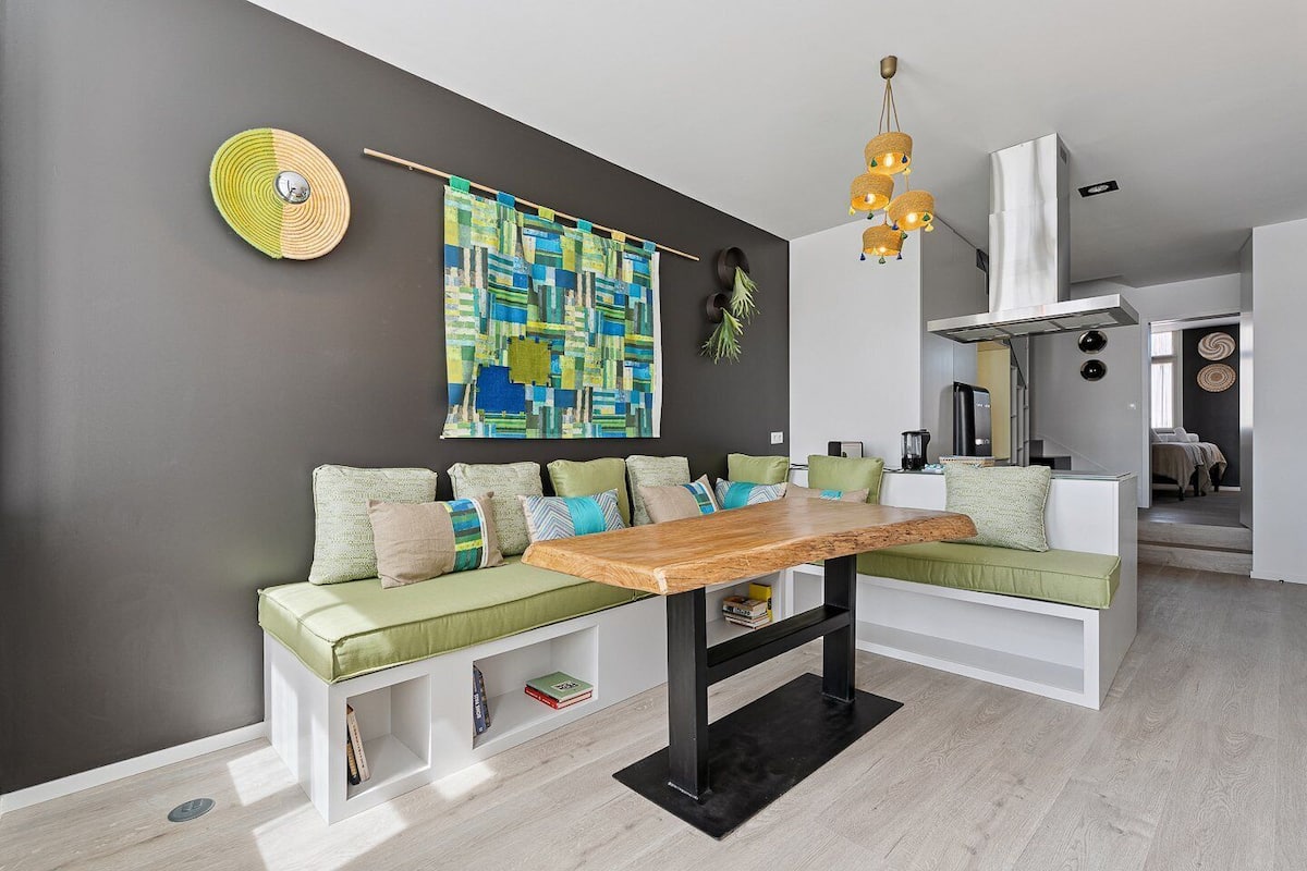 Ein modernes Wohnzimmer mit einem Holztisch, grün-beigen Kissen, einem farbenfrohen abstrakten Gemälde und einer außergewöhnlichen Deckenleuchte.
