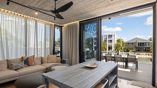 A modern living room with a sofa, a dining table, and a view of a balcony with outdoor furniture. - Oatland 26 Esplanade Coastal Retreat (Runaway Bay)