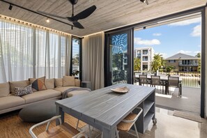 A modern living room with a sofa, a dining table, and a view of a balcony with outdoor furniture. - Oatland 26 Esplanade Coastal Retreat (Runaway Bay)