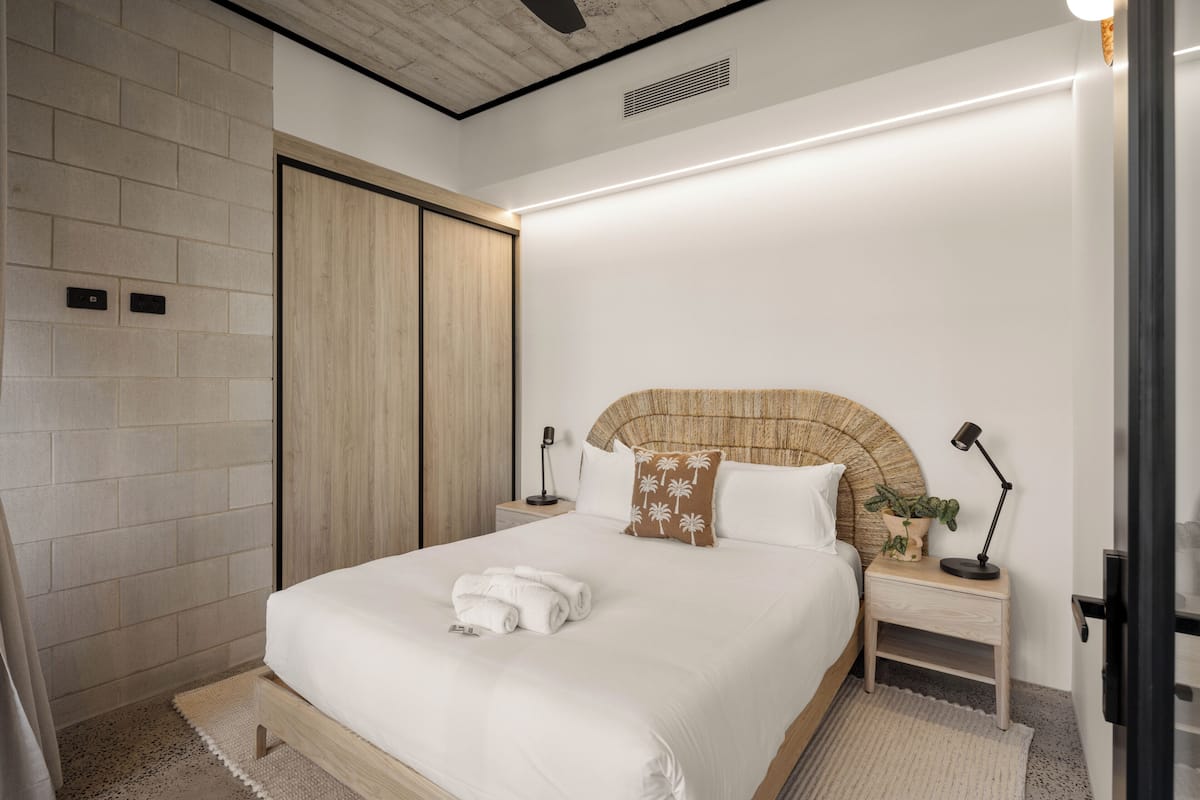 Une chambre d’hôtel moderne avec un grand lit, une tête de lit en bois, des tables de chevet avec des lampes, une plante en pot et une armoire intégrée.