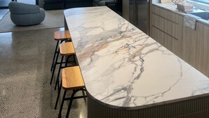 A modern kitchen with a marble countertop, bar stools, a built-in oven, and a large window overlooking a balcony. - Oatland 26 Esplanade Coastal Retreat (Runaway Bay)