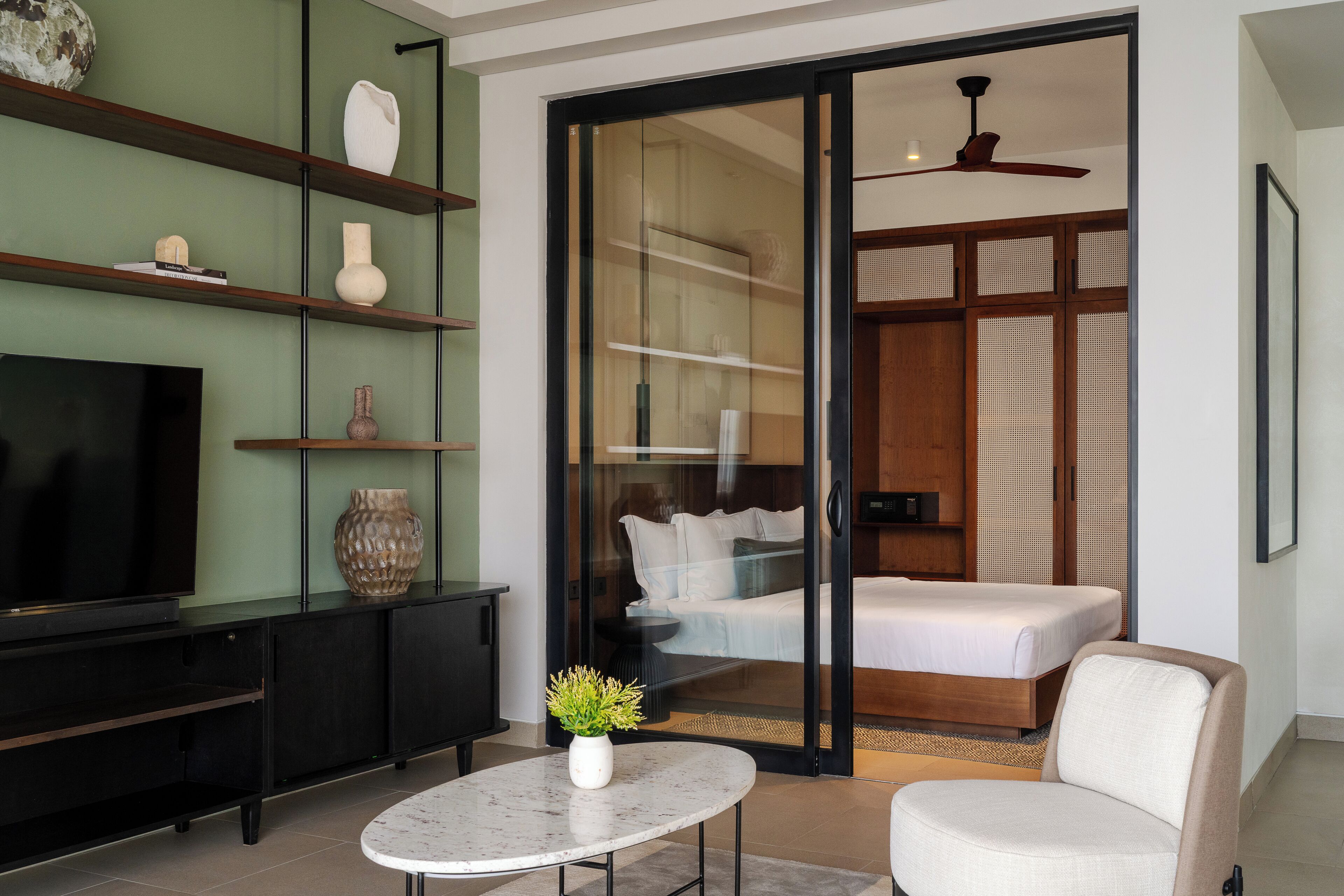 A modern hotel room with a flat-screen TV, a marble coffee table, a sofa, a bed with a white bedspread, and a sliding glass door leading to a balcony.