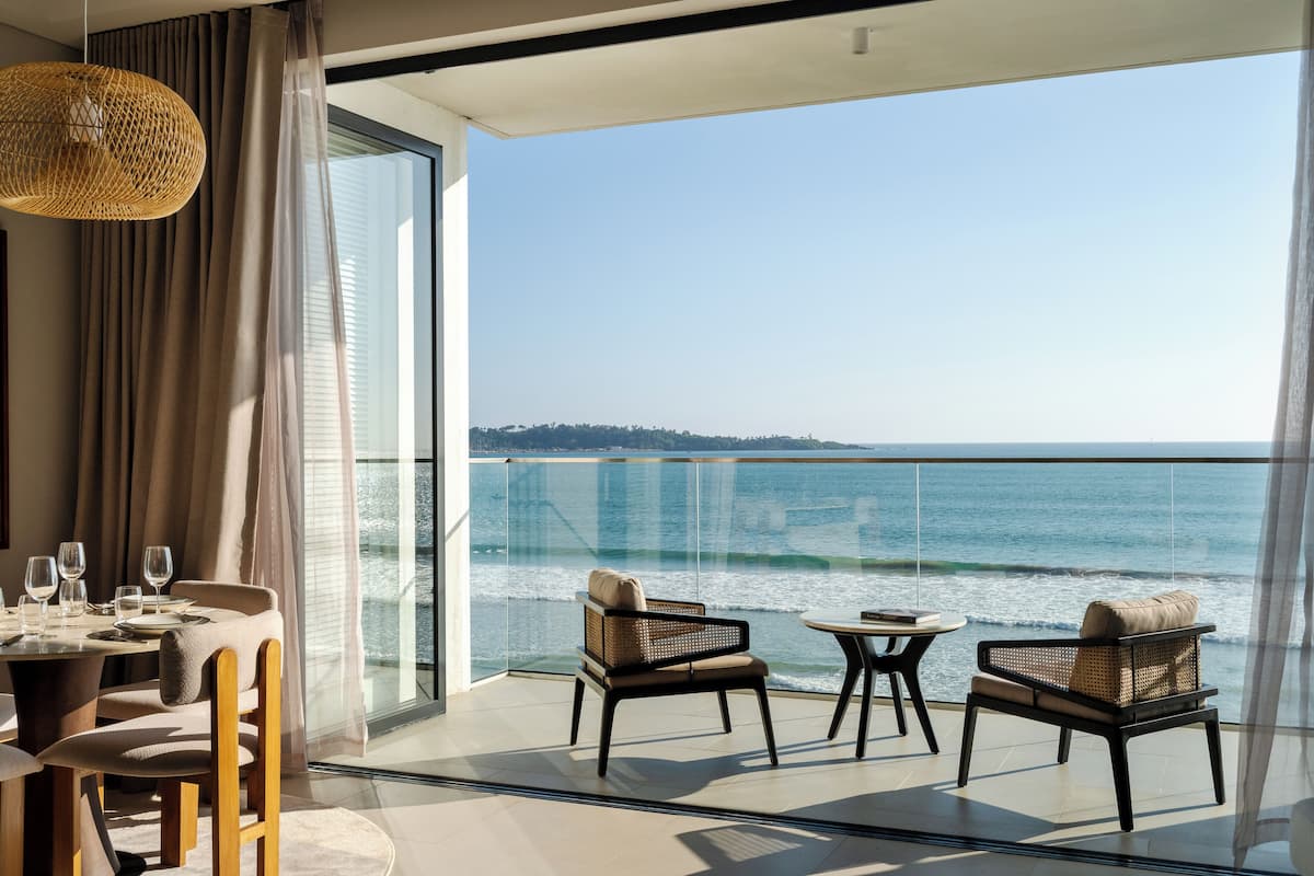 Un balcon avec vue sur la mer, des fauteuils en osier, une table à manger dressée pour deux personnes et un luminaire suspendu.