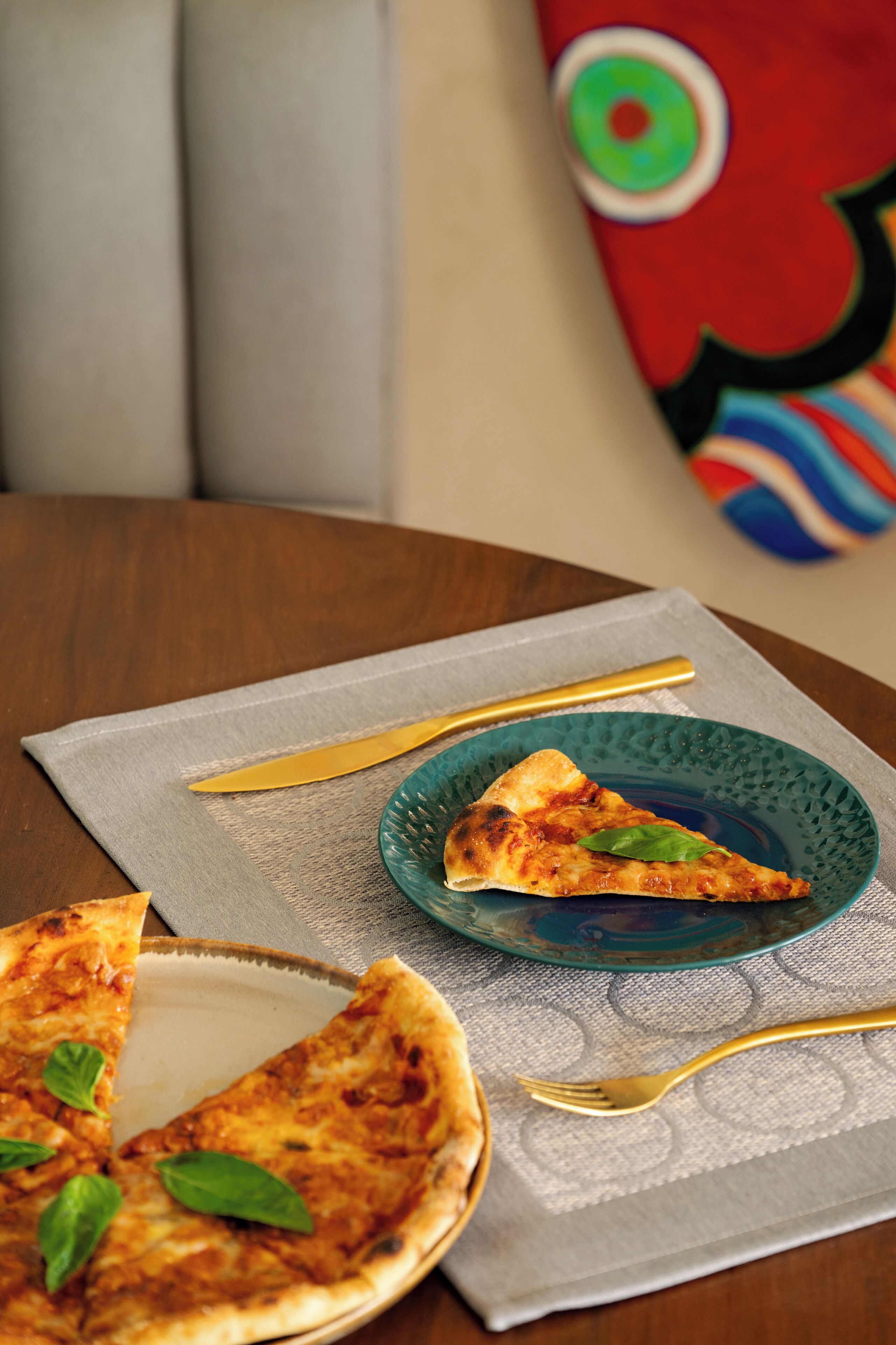 A pizza on a blue plate with a slice missing, a fork, and a knife on a tablecloth.