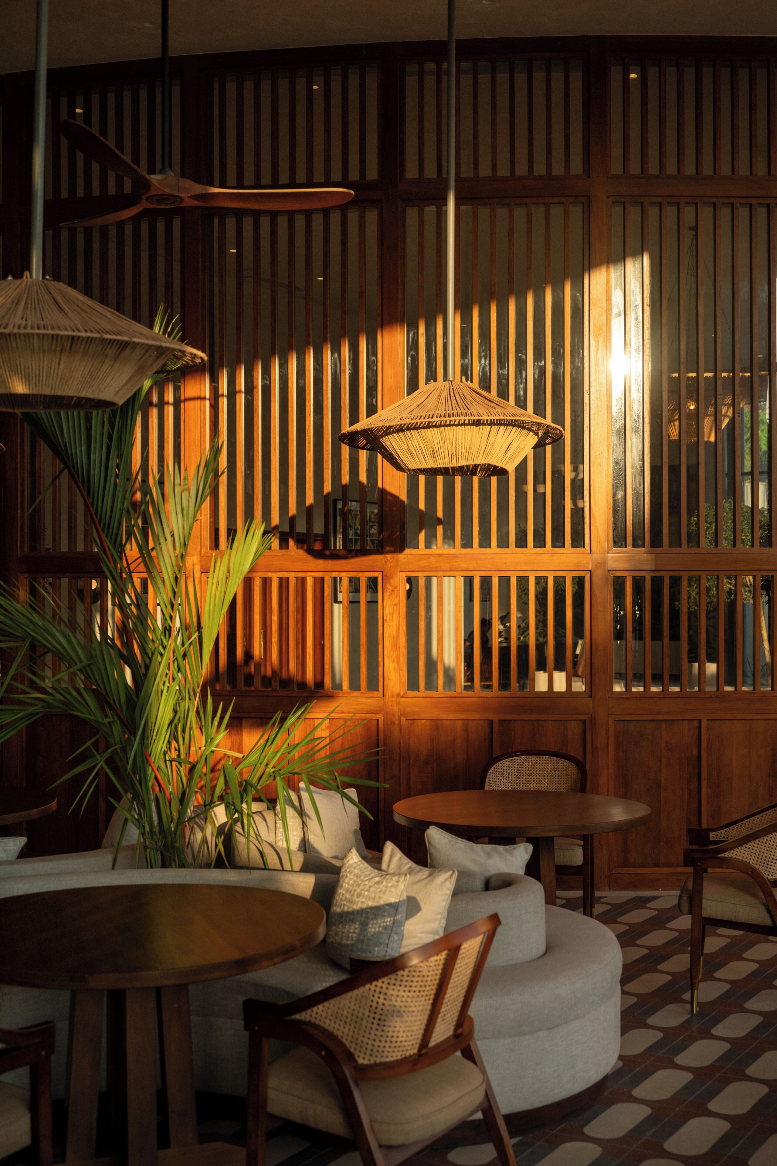 A room with a wooden partition, round tables, cushioned seating, and woven ceiling lamps.