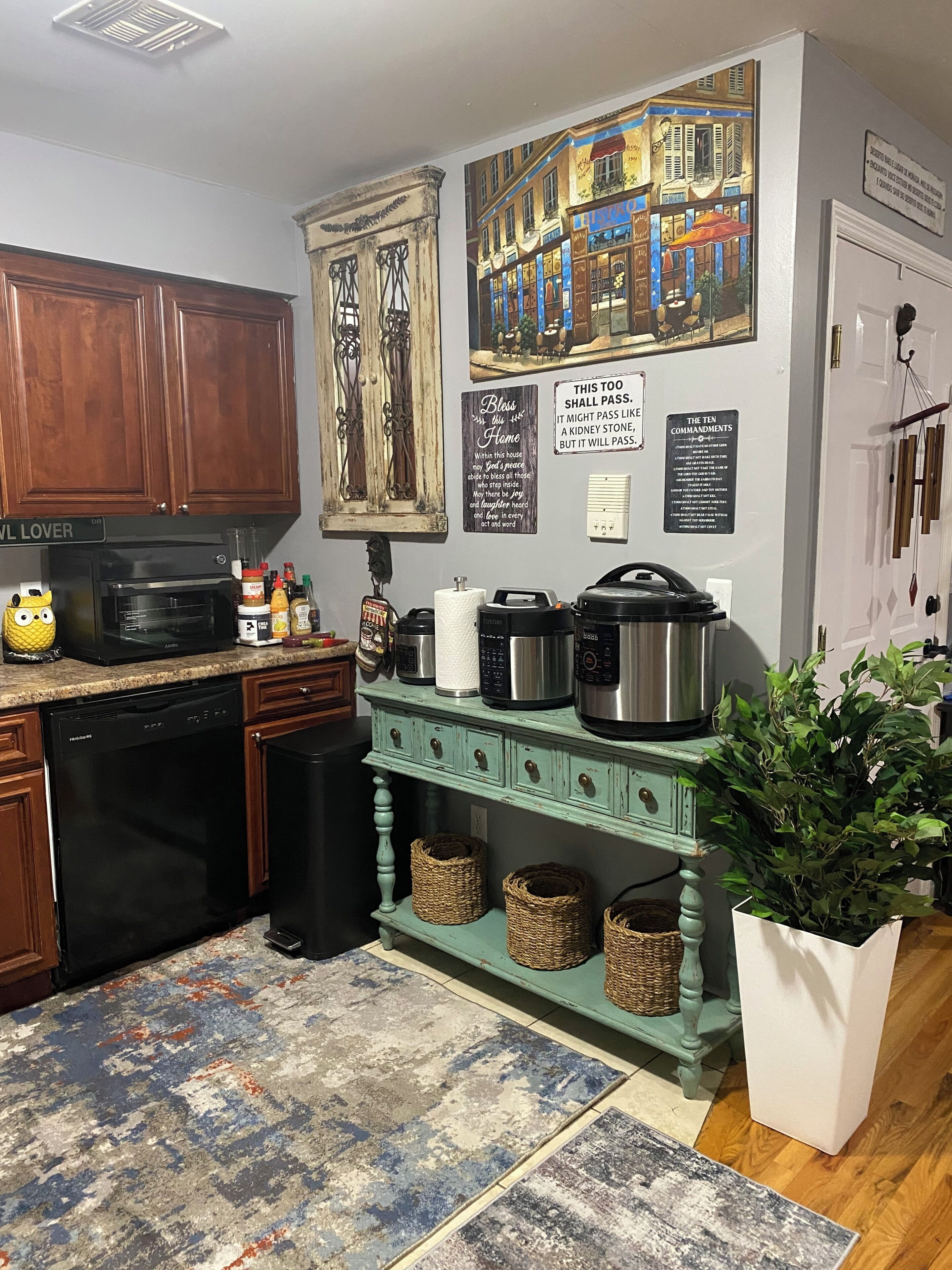 A kitchen with wooden cabinets, a dishwasher, a microwave, a coffee maker, and a kettle. There are wicker baskets on a green cabinet, a potted plant, and a painting of a street scene on the wall.