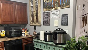 Une cuisine avec des meubles en bois, un lave-vaisselle, un four à micro-ondes, une machine à café et une bouilloire. On y trouve des paniers en osier sur un meuble vert, une plante en pot et un tableau représentant une scène de rue accroché au mur.