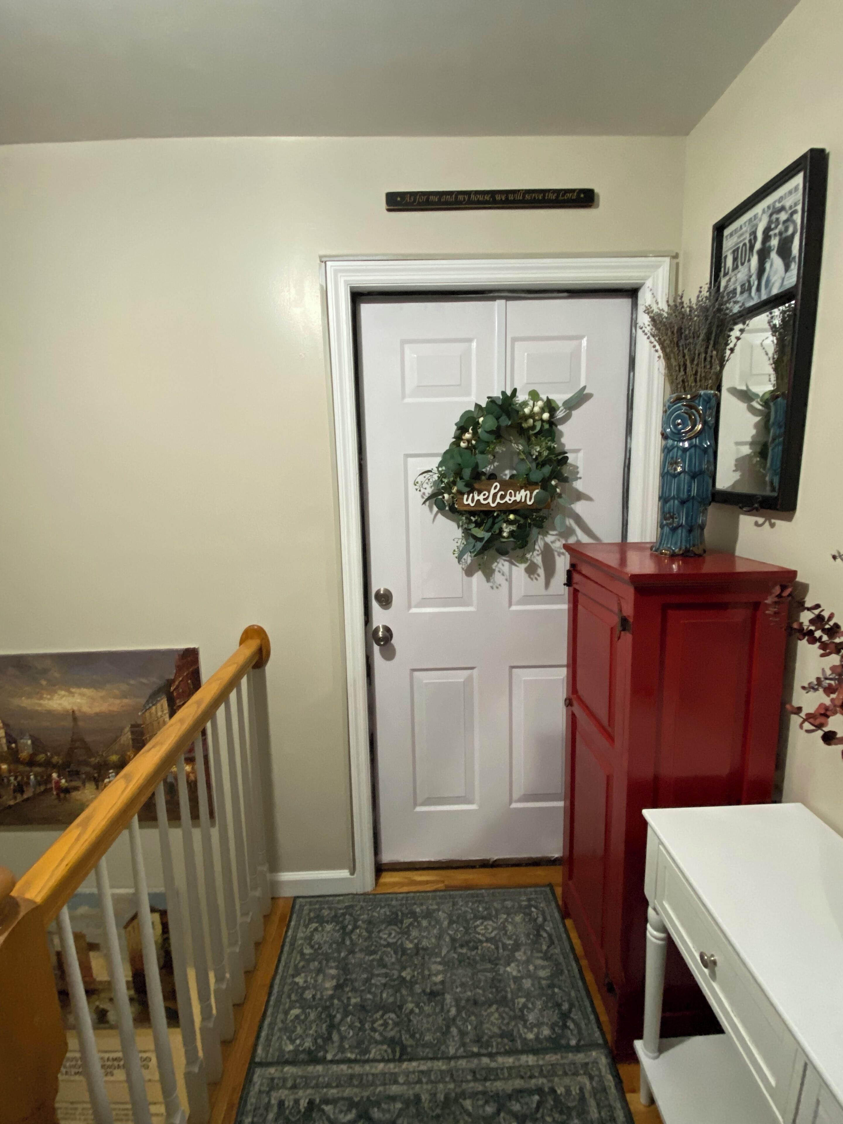 Uma porta branca com uma grinalda e um letreiro de 'welcome', um armário vermelho, um quadro emoldurado e um lance de escadas de madeira.