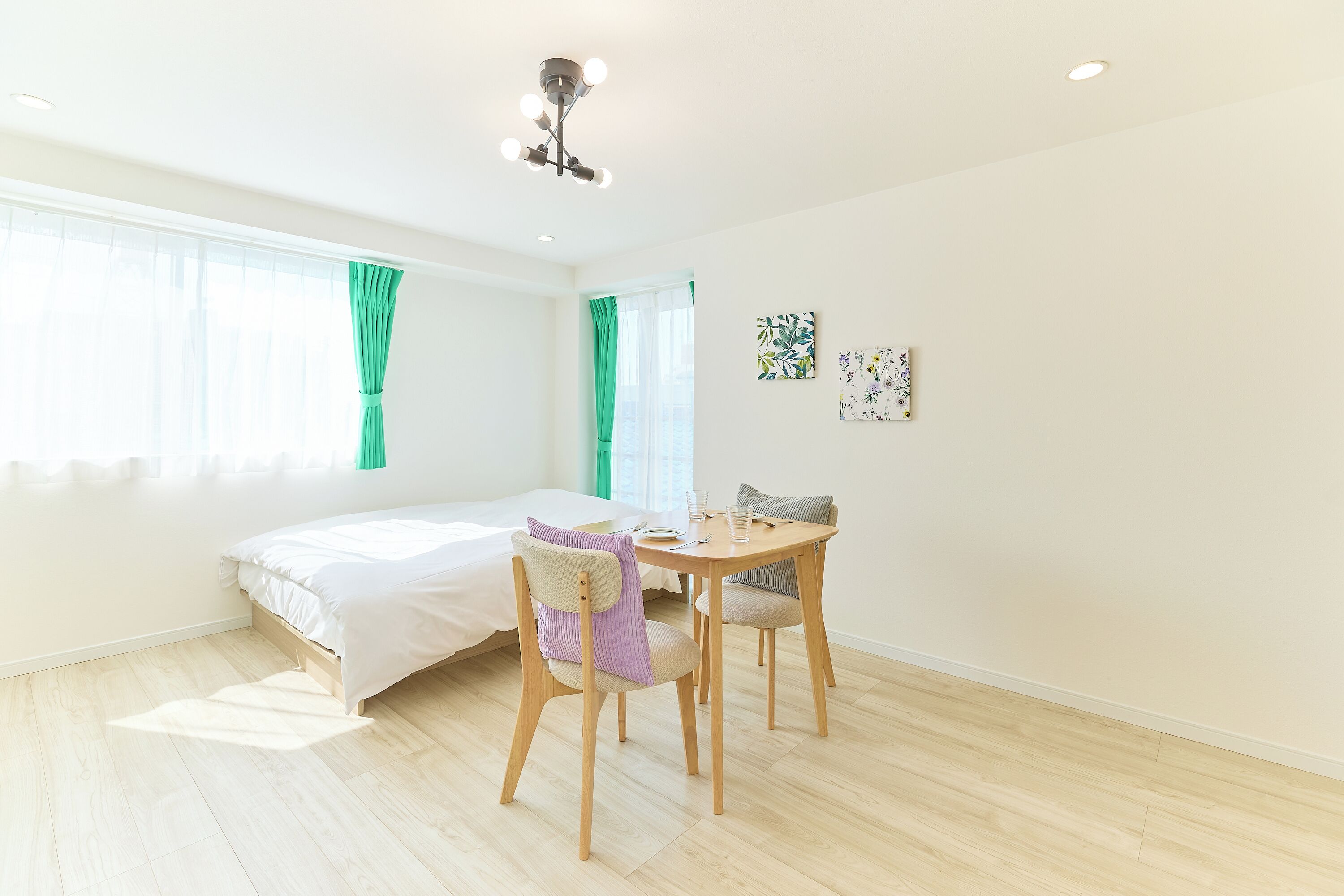 A room with a bed, a dining table with chairs, a ceiling light, and two windows with curtains.