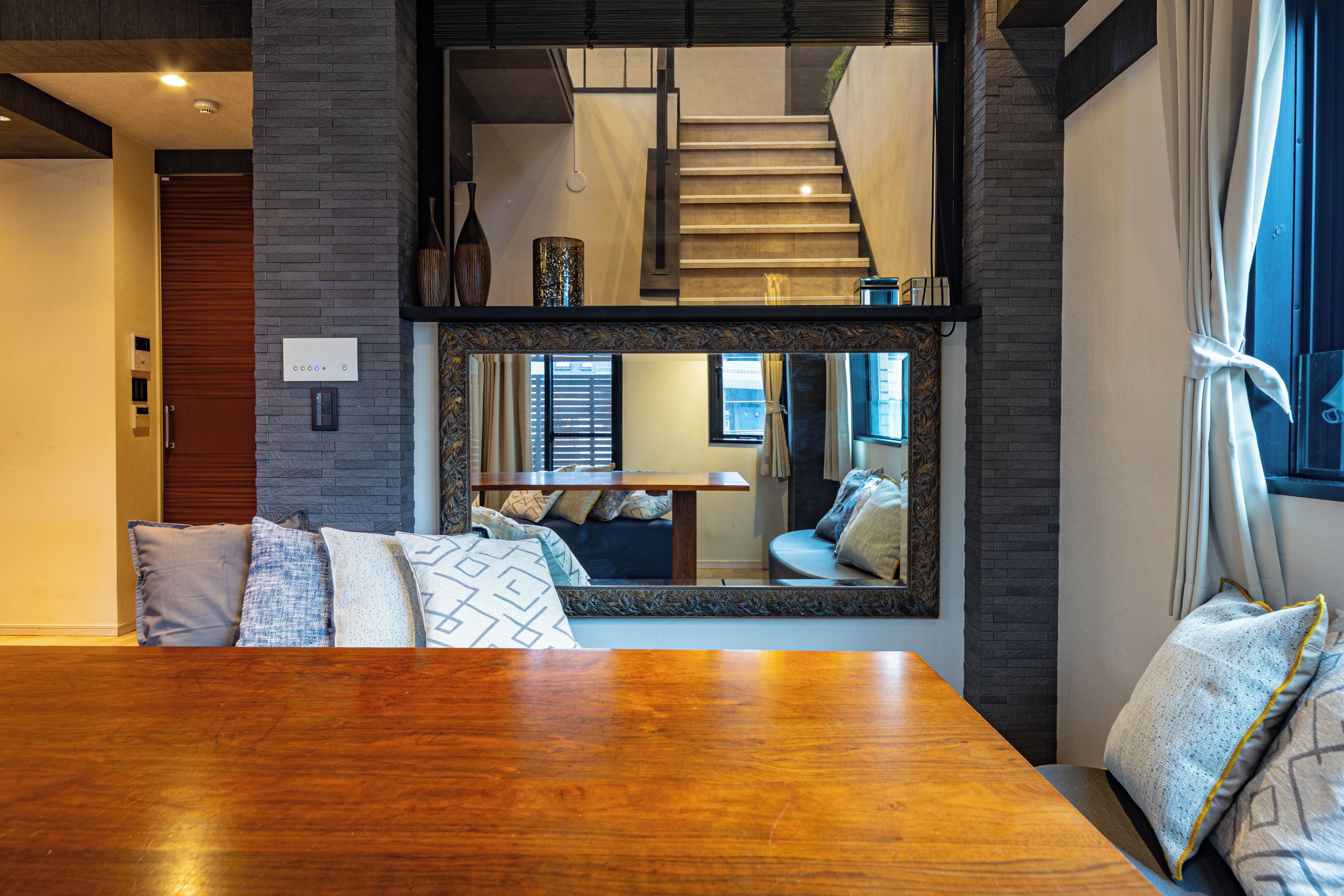 A modern interior with a wooden dining table, a large mirror reflecting a living area, and a staircase leading to an upper level.
