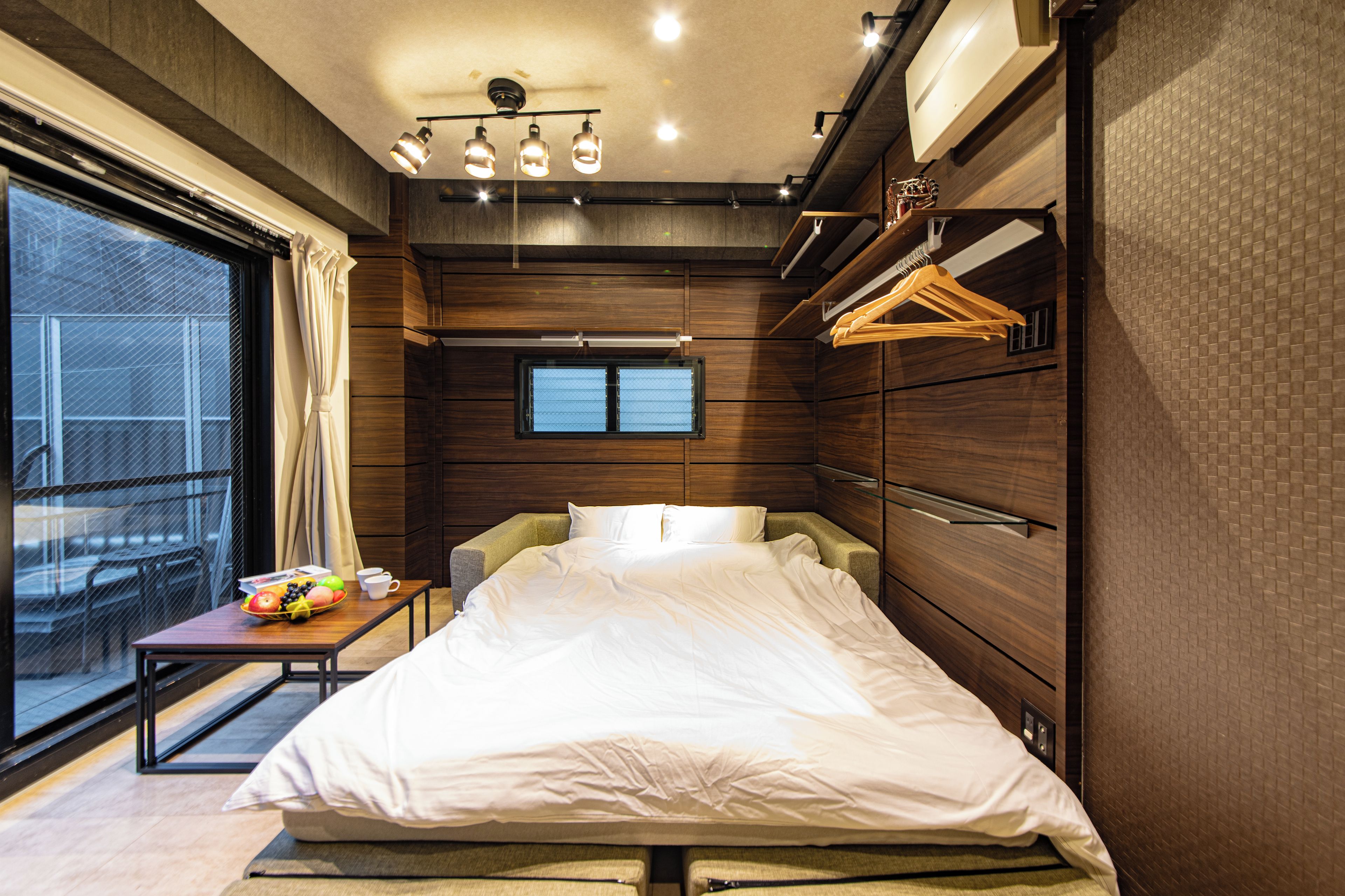 A modern bedroom with a large bed, a small table with a fruit bowl, a window, and a wardrobe with hanging clothes.