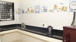 A kitchen with white tiled backsplash, black countertop, stainless steel appliances, and a window with a metal grill. - Jayam Residency (Tiruvannamalai)