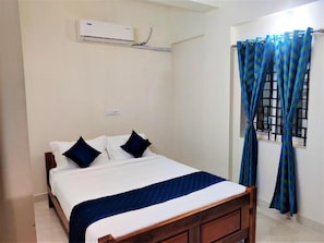 A single bed with a wooden frame, a white mattress, and a blue quilt. There is a wall-mounted air conditioner above the bed.