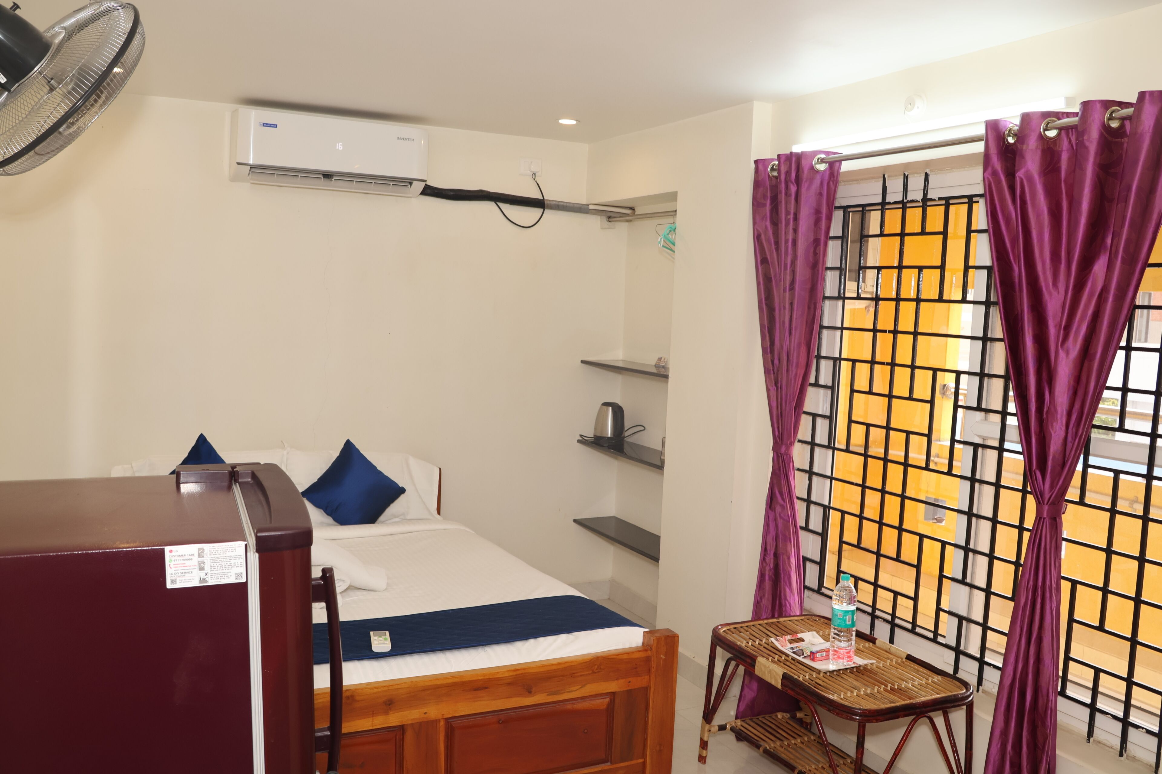 Habitación de hotel con cama, una mesita, un ventilador, una tetera y una ventana con cortinas moradas.
