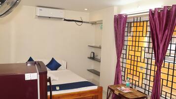 Habitación de hotel con cama, una mesita, un ventilador, una tetera y una ventana con cortinas moradas.