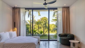 A bedroom with a bed, a green chair, a small table, and a view of greenery through large windows.