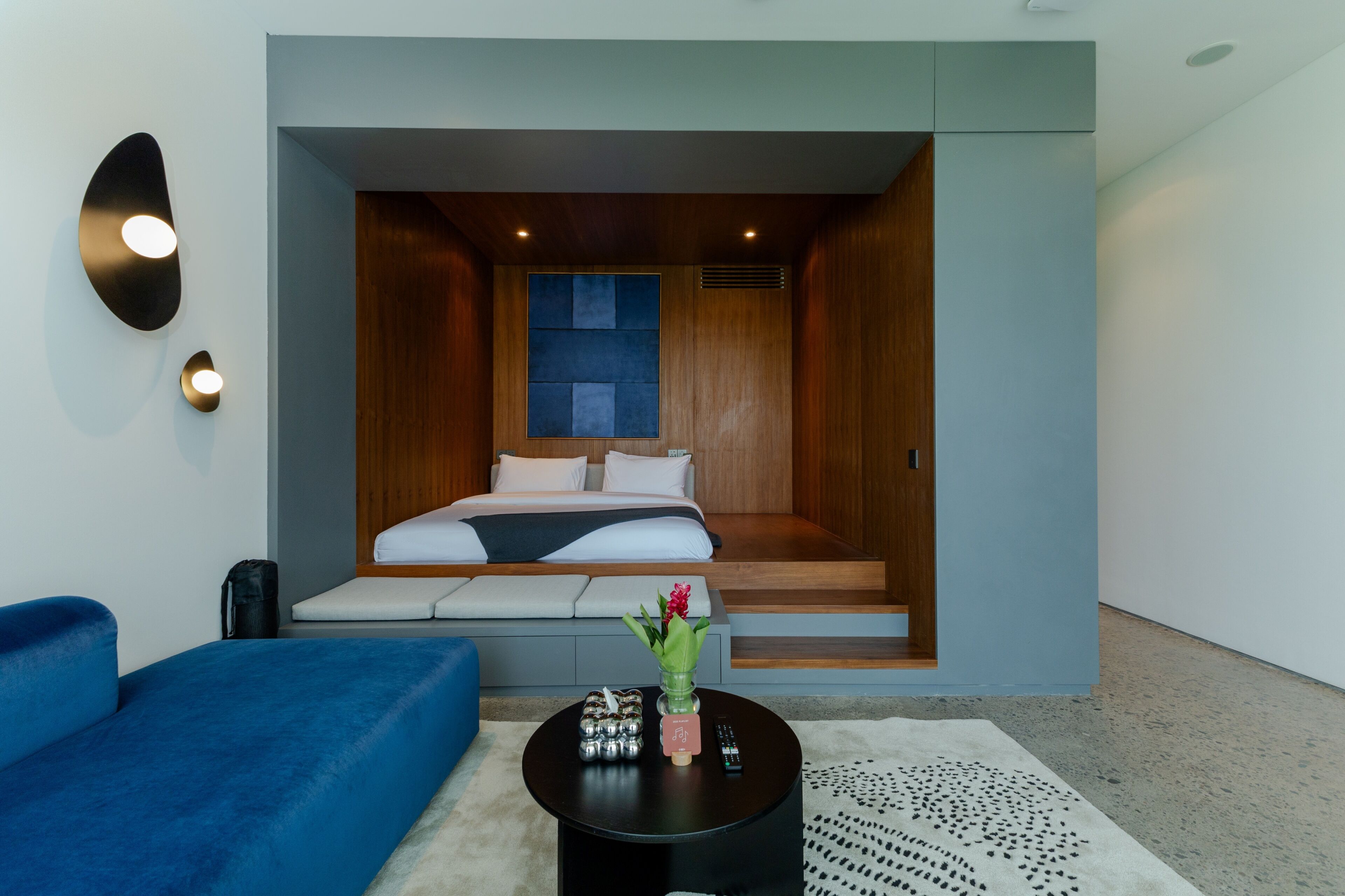 A modern hotel room with a bed, a blue sofa, a small table with a vase, and a wall-mounted light fixture.