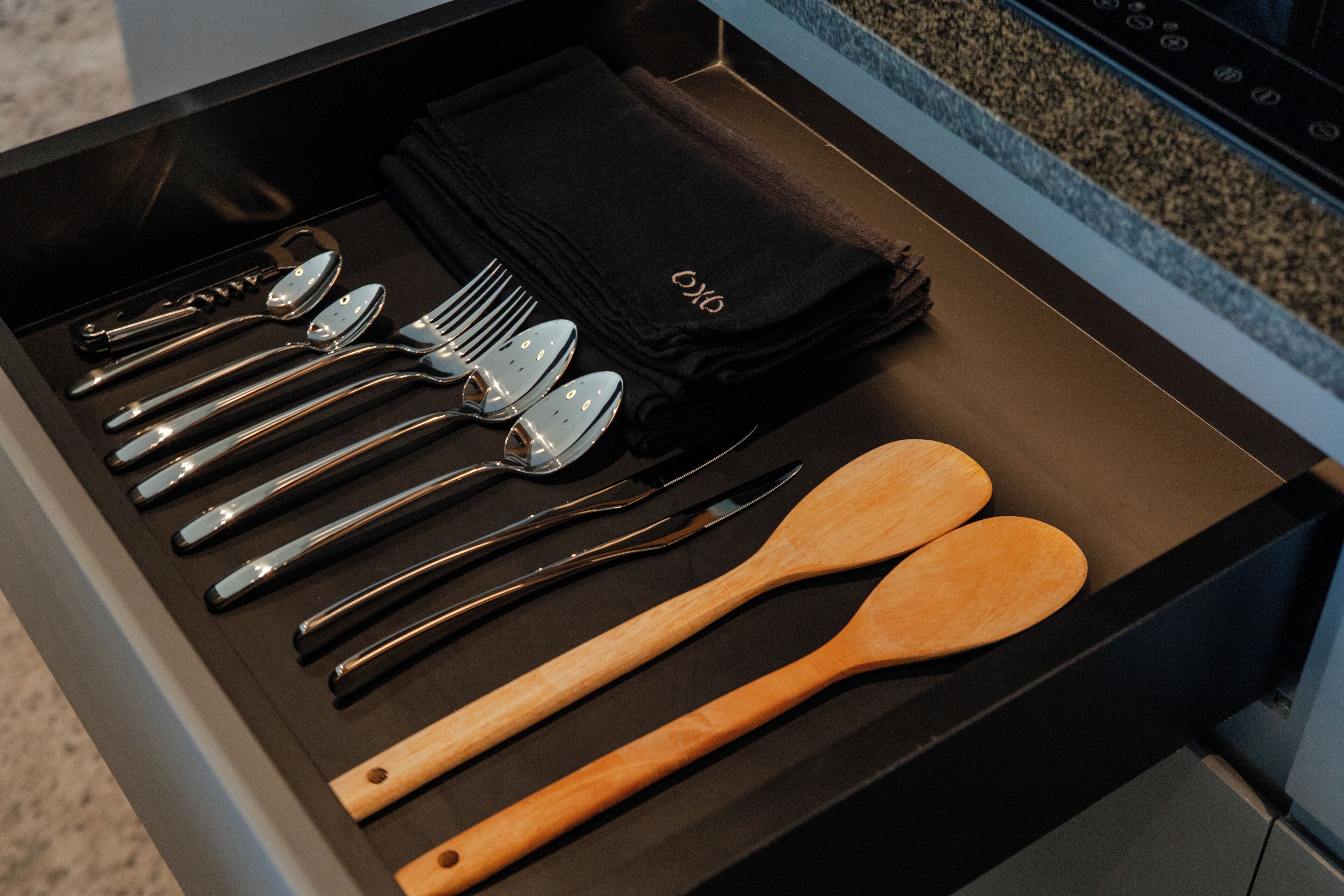 Cajón con interior negro que contiene cubiertos, cucharas de madera y un paño negro con la marca 'OXO'.