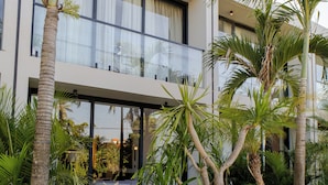 A modern building with large windows, a balcony, and a garden with palm trees and a wooden deck.