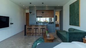 A modern hotel room with a kitchenette, a dining area with bar stools, a green sofa, a TV, and a large artwork on the wall.