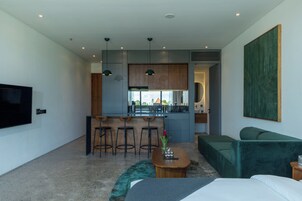 A modern hotel room with a kitchenette, a dining area with bar stools, a green sofa, a TV, and a large artwork on the wall.